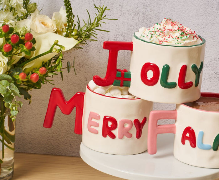 The Sophistiplate Ceramic Message Mug "Merry," 14oz, features a festive design with colorful letters and is perfect for enjoying your favorite holiday drinks. A vase with white flowers and greenery sits to the left.