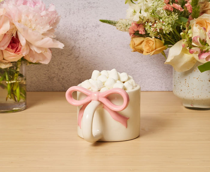 The Sophistiplate Ceramic Pink Bow on Handle Mug, 12oz, filled with mini marshmallows, rests on wood. Flower arrangements in glass and ceramic vases decorate the background against a light wall. Microwave safe.