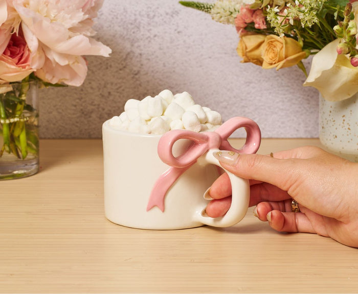 A hand holds the Sophistiplate Ceramic Pink Bow on Handle Mug, 12oz, filled with mini marshmallows. The mug rests on a wooden table, with vases of assorted flowers in the background.