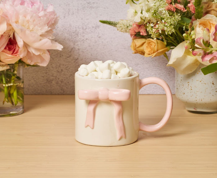 A cream-colored Ceramic Pink Bow Side Mug, 16oz by Sophistiplate, microwave and dishwasher safe, filled with mini marshmallows sits on a wooden surface. Soft-hued flower vases decorate the background.