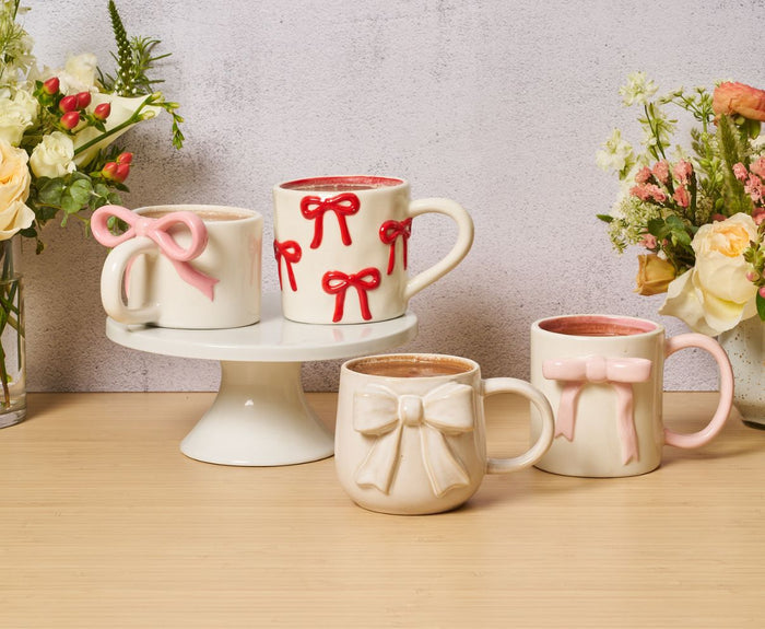 Four Sophistiplate Ceramic Pink Bow Side Mugs (16oz) are arranged on a wooden surface, some on a cake stand with fresh flowers. Each mug is microwave and dishwasher safe for daily convenience.