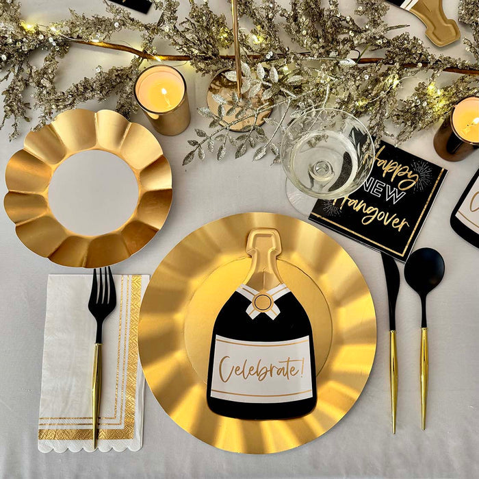 The Sophistiplate Champagne Celebration Table Set features designer disposable plates, black cutlery, gold-trimmed napkins, a “Happy New Year” napkin, candles, and a champagne bottle-shaped Celebrate! card on a white tablecloth with festive décor.