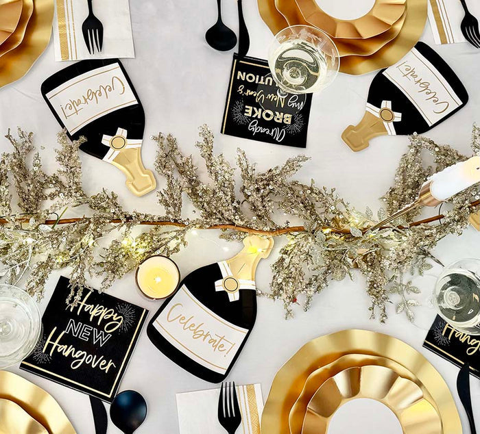 Create a festive table with the Champagne Celebration Table Set by Sophistiplate—includes gold designer disposables, black cutlery, champagne bottle decorations, themed napkins ("Celebrate", "Happy New Hangover", "Break Tradition"), and a gold garland centerpiece.
