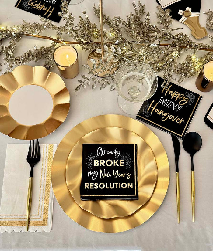 Create a festive table with the Sophistiplate Champagne Celebration Table Set featuring designer gold plates, black and gold cutlery, a candle, gold accents, and black napkins with witty New Year’s sayings.