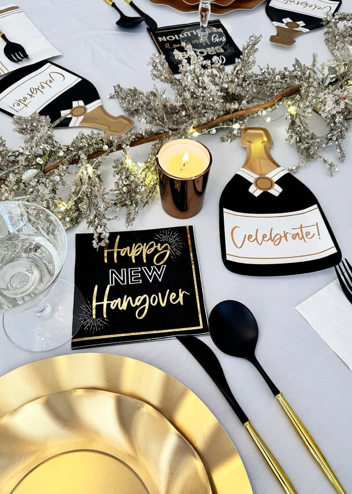 Set a festive scene with the Sophistiplate Champagne Celebration Table Set, featuring designer disposable plates, gold and black cutlery, a lit candle, sparkling branch centerpiece, and New Year decor like "Happy New Hangover" napkins.