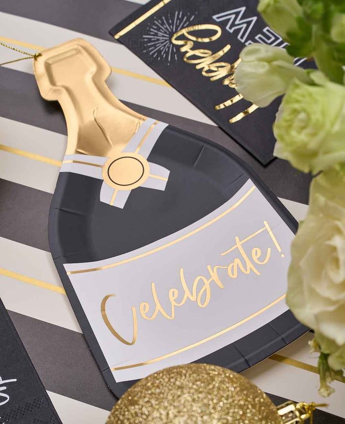 The Sophistiplate Champagne Celebration Table Set features a champagne bottle-shaped paper decoration with "Celebrate!" in gold, white flowers, festive black cards, gold ornaments, and stylish designer plates on a striped surface.