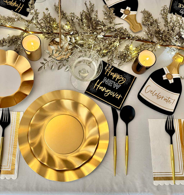 Create a festive table with the Sophistiplate Champagne Celebration Table Set, featuring gold disposable plates, black and gold cutlery, white napkins, candles, and New Year's decorations like “Happy New Hangover” signs and champagne bottle cutouts.