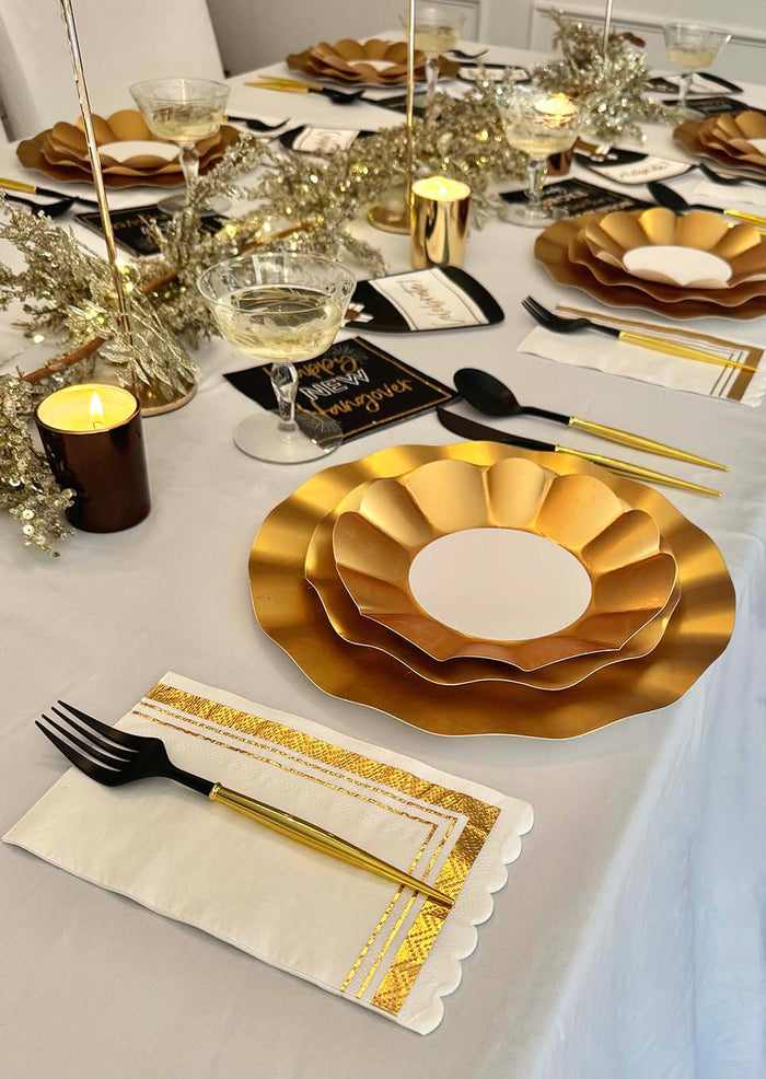 The Champagne Celebration Table Set by Sophistiplate features gold scalloped plates, gold flatware, black forks, champagne glasses, white napkins with gold trim, candles, and gold garland on a white tablecloth.