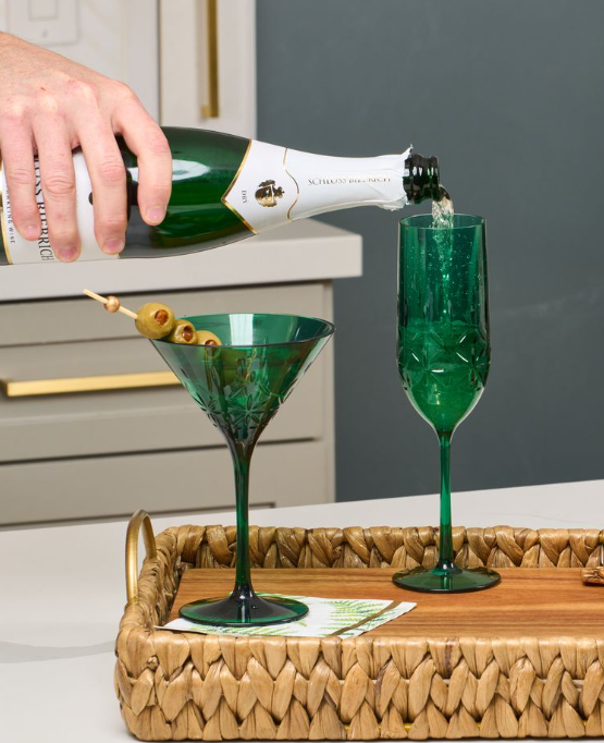 A hand pours sparkling wine into a green Sophistiplate Champagne Classic 8.9 Oz flute on a wooden tray, next to a matching green martini glass with olives. Both are BPA-free plastic and dishwasher safe for easy cleanup.
