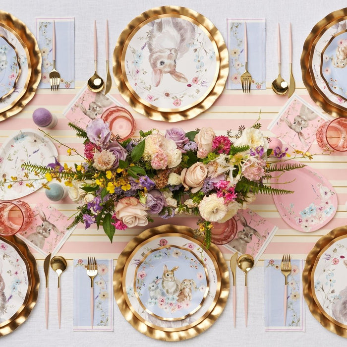 Create a festive look with the Sophistiplate Charming Easter Table Setting, featuring bunny and chick plates, gold cutlery, pink and purple glassware, striped napkins, plus a floral centerpiece with eggs and greenery.