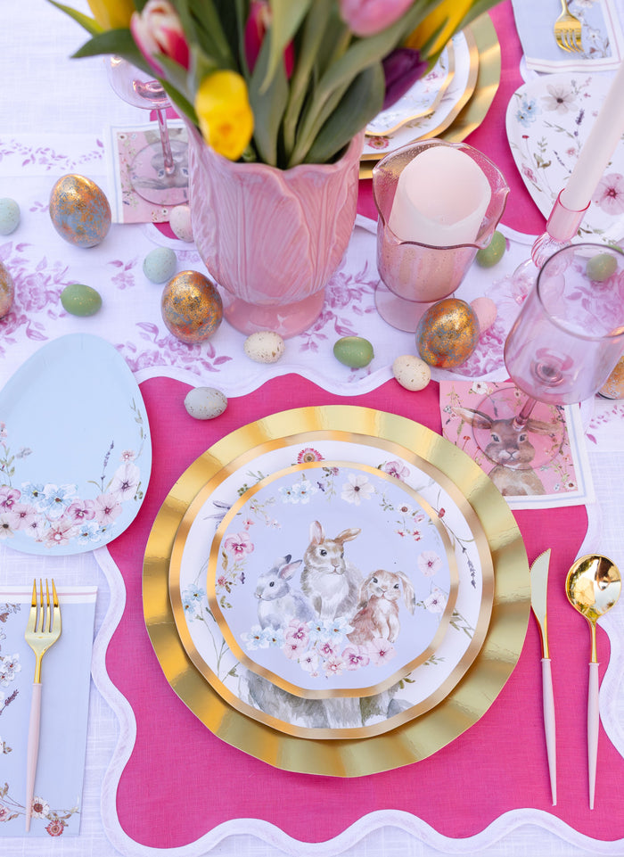 Create a festive Easter table for 16 with Sophistiplate LLC’s Ultimate Easter Tablescape: Charming Easter—designer bunny plates, gold-rimmed chargers, pink and white linens, gold cutlery, pastel eggs, tulips in a pink vase, candles, and decorative napkins.