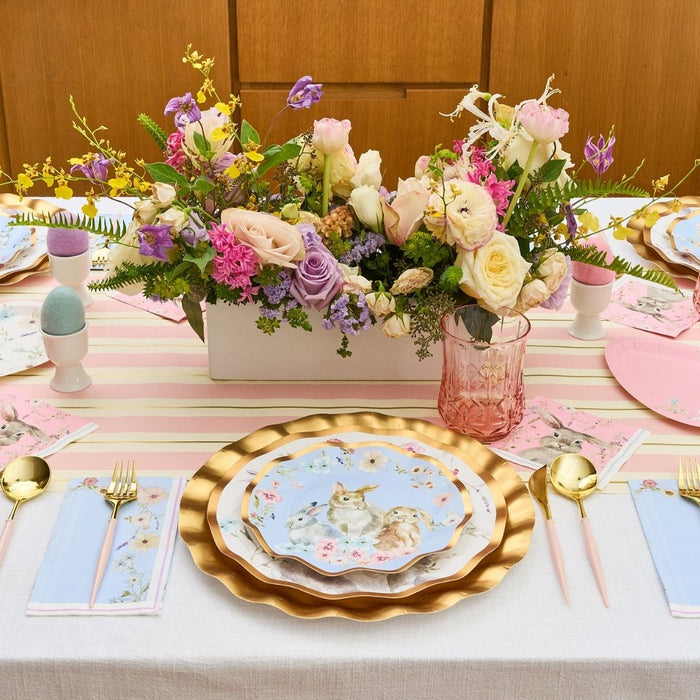 The Charming Easter Table Setting by Sophistiplate features bunny-illustrated plates, pink and gold cutlery, pastel napkins, colored eggs in egg cups, and a floral centerpiece with roses and wildflowers for a festive spring look.
