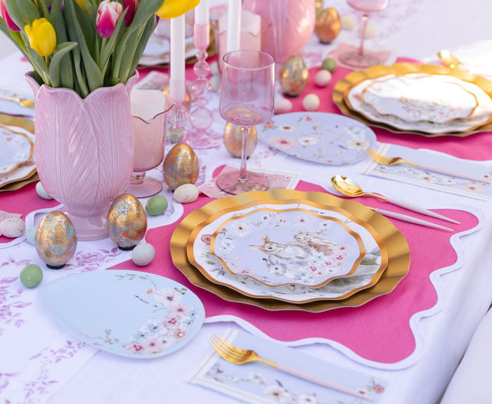 Create a pastel-themed Easter table with Sophistiplate LLC’s Ultimate Easter Tablescape for 16: Charming Easter, featuring pink tulips, gold and pink cutlery, rabbit paper plates, gold eggs, pink glasses, and white candles on a festive cloth.