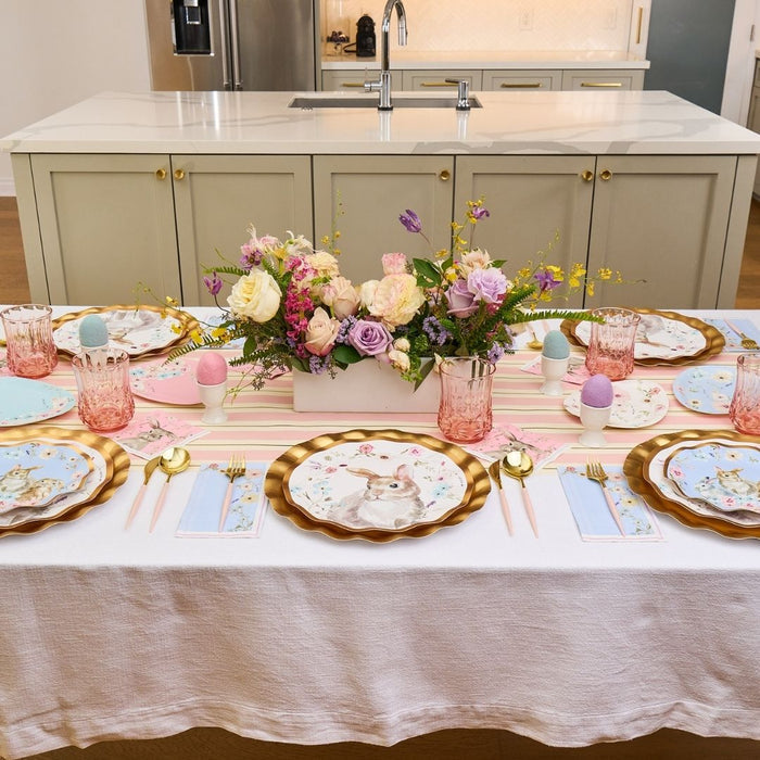 The Charming Easter Table Setting by Sophistiplate features pastel bunny plates, gold-rimmed chargers, pink glasses, and Easter egg decor with designer paper plates and a floral centerpiece for a festive tablescape.