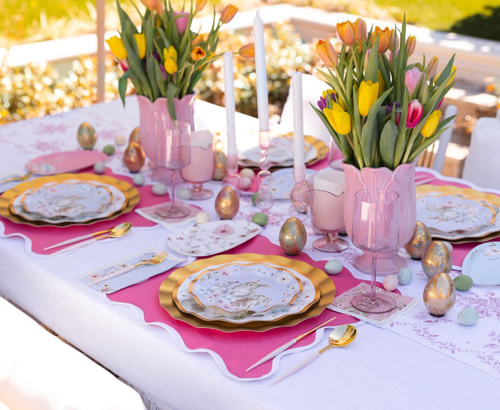 Create a spring-inspired Easter table with Sophistiplate LLC's Ultimate Easter Tablescape for 16: Charming Easter—featuring gold-rimmed designer plates, pink glasses, gold cutlery, tulips, pastel candles, and decorative eggs on a chic white cloth.