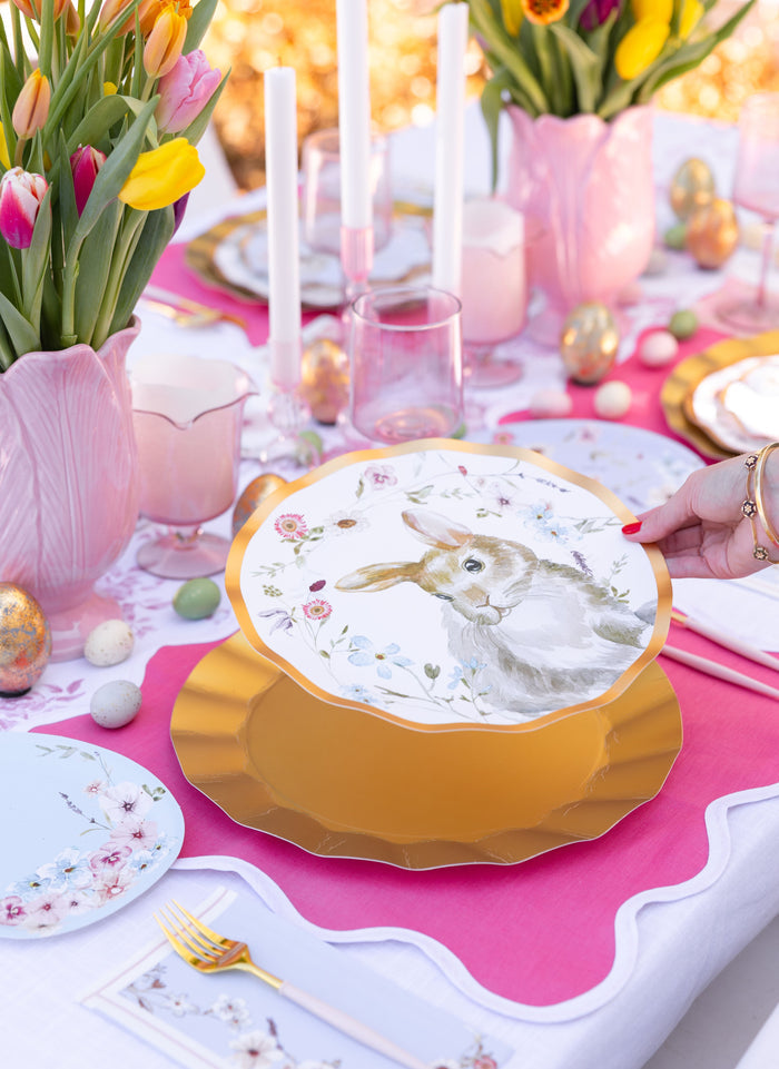 The Ultimate Easter Tablescape for 16: Charming Easter by Sophistiplate LLC features pink and gold accents, tulip arrangements, candles, pastel eggs, and designer rabbit plates held above a gold charger for a stylish spring look.
