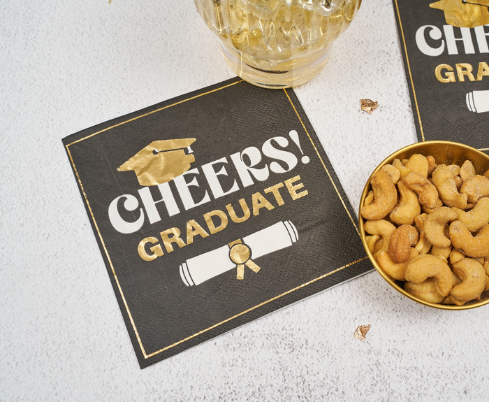 Celebrate with Sugarplum Cheers Graduate Cocktail Napkins (26pk)! These black napkins feature gold and white “CHEERS! GRADUATE” text with a cap and diploma—perfect for your graduation party snacks and drinks.