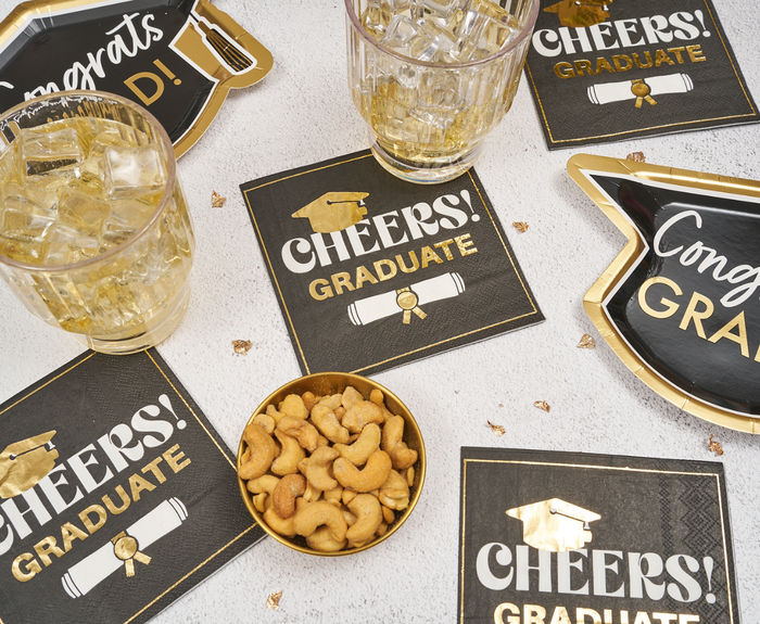 Sugarplum Cheers Graduate Cocktail Napkins (26pk) are set on a table with two glasses of ice, a bowl of cashews, and scattered confetti, creating a festive graduation party display.