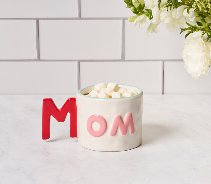 The Cheers to Mom Gift Set 2 by Sophistiplate LLC features a white mug with "MOM" in red and pink, filled with mini marshmallows on a marble countertop—an ideal Mother’s Day gift. White flowers appear in the top right against tiled walls.