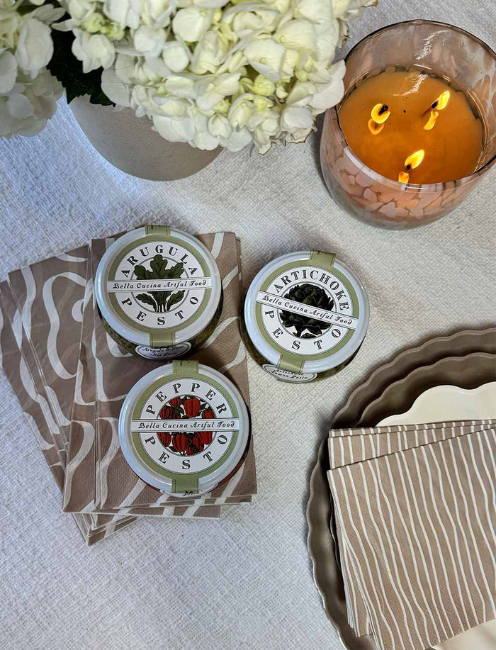 The Chef’s Selection Pesto Set from Bella Cucina sits on patterned brown napkins beside stacked plates, a striped cloth, white hydrangeas, and a lit candle—ideal for elevating your next charcuterie board.