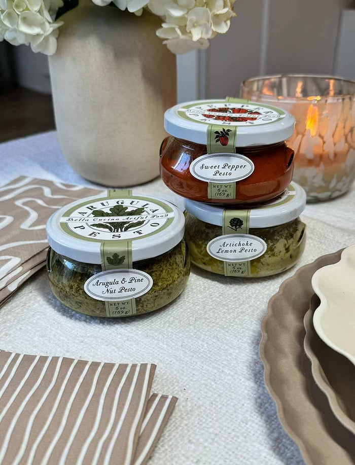 Three jars from the Bella Cucina Chef’s Selection Pesto Set are stacked on a table, ready to enhance your charcuterie board. Plates, napkins, a lit candle, and white flowers complete this inviting gourmet scene.