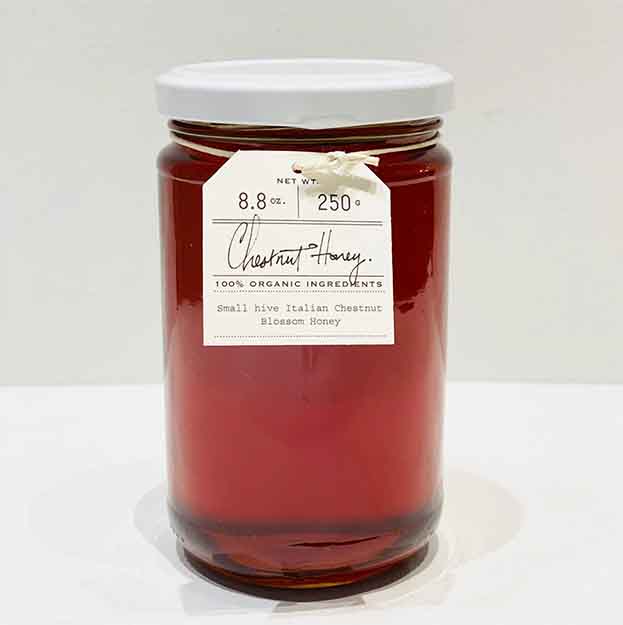 A glass jar with a white lid contains Bella Cucina Chestnut Honey—amber in color, 100% organic, 8.8 oz (250g), and authentic biodynamic honey from Piedmont, Italy.
