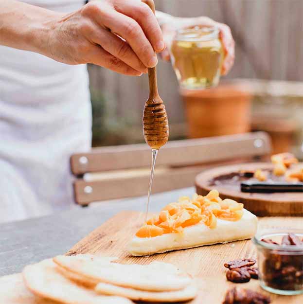 A person drizzles Bella Cucina Organic Acacia Honey over cheese topped with chopped dried apricots on a wooden board, surrounded by crackers, nuts, and a glass of white wine. This organic honey from Piedmont, Italy imparts a delicate floral sweetness to the elegant spread.