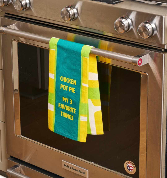 A colorful Chicken Pot Pie Dish Towel by Drinks On Me hangs on a stainless steel oven handle. This humorous kitchen towel features the phrase "CHICKEN POT PIE MY 3 FAVORITE THINGS" in yellow letters.