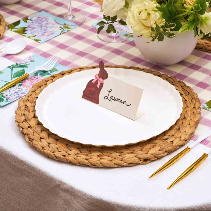 The Ultimate Easter Tablescape for 16: Violet Hydrangea by Sophistiplate LLC features woven placemats, designer disposable plates, gold cutlery, floral napkins, bunny name card holders, and a floral centerpiece on a pink plaid tablecloth.