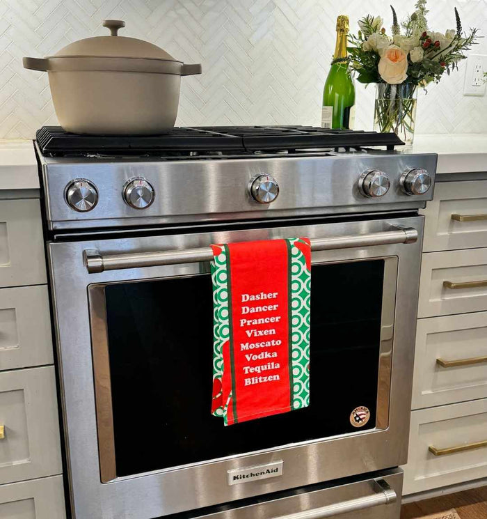 A modern stainless steel oven displays the CHRISTMAS Blitzen Dish Towel by Drinks On Me, listing reindeer names and drinks for a humorous touch. With flowers, a pot, and champagne on the white countertop, it's perfect kitchen humor for housewarming gifts.