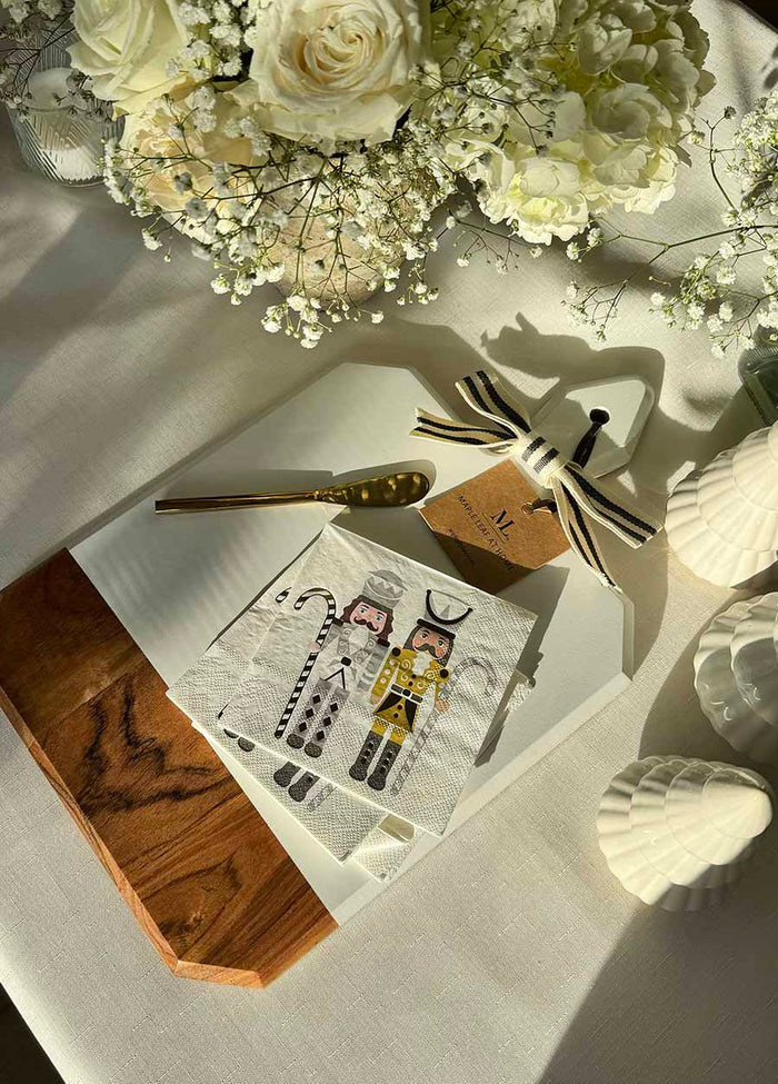 The Sophistiplate Christmas Board Gift Kit: Gilded Nutcracker features a festive notepad with pen, chef and waiter napkin, ribbon, white flowers, all artfully set on an acacia wood charcuterie board—a perfect holiday hostess gift.