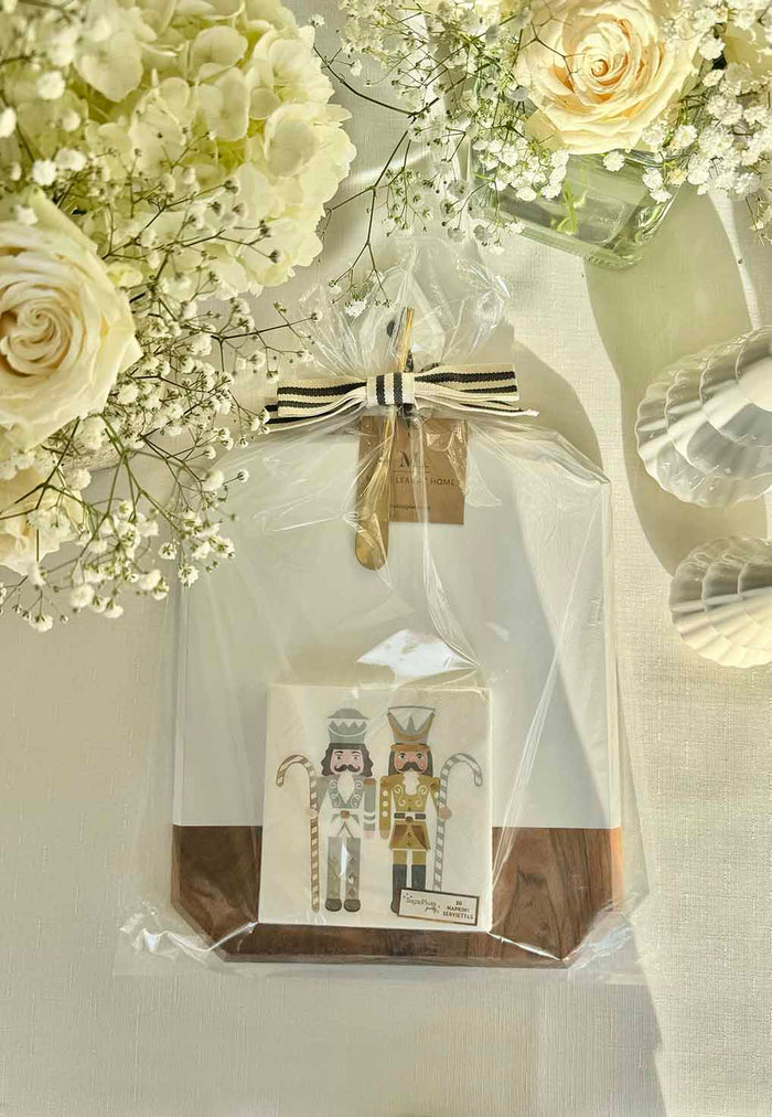 The Sophistiplate Christmas Board Gift Kit: Gilded Nutcracker is wrapped in clear plastic with a black and white striped ribbon, surrounded by white roses and baby’s breath. A card featuring two illustrated nutcrackers is attached to the front.