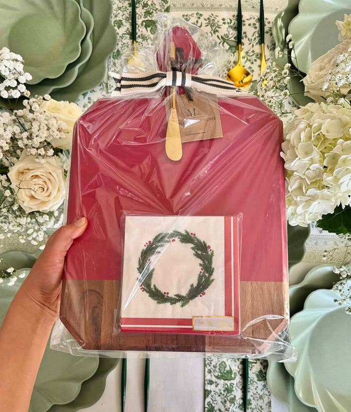A hand holds the Sophistiplate Christmas Board Gift Kit: Merry & Bright, featuring a red wrap, wooden base, and holiday wreath napkin. White flowers and green floral tableware create a festive background.