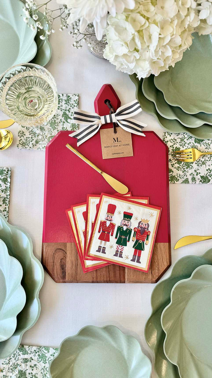 A festive table features Sophistiplate's Christmas Board Gift Kit: Nutcracker, with nutcracker-themed napkins, a gold spreader, and ribbon, paired with pale green scalloped plates, floral napkins, a wine glass, and an acacia charcuterie board.