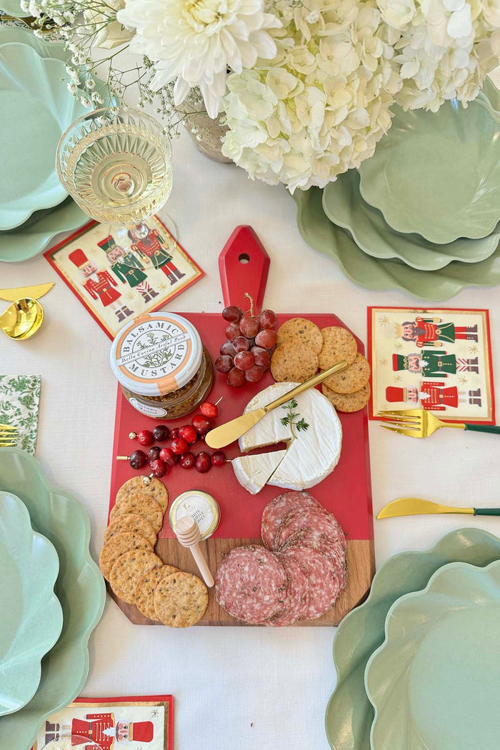 The Sophistiplate Christmas Board Gift Kit: Nutcracker displays brie, crackers, salami, grapes, cranberries, and jam on an acacia wood and red platter with green scalloped plates, nutcracker napkins, gold flatware, and white flowers.