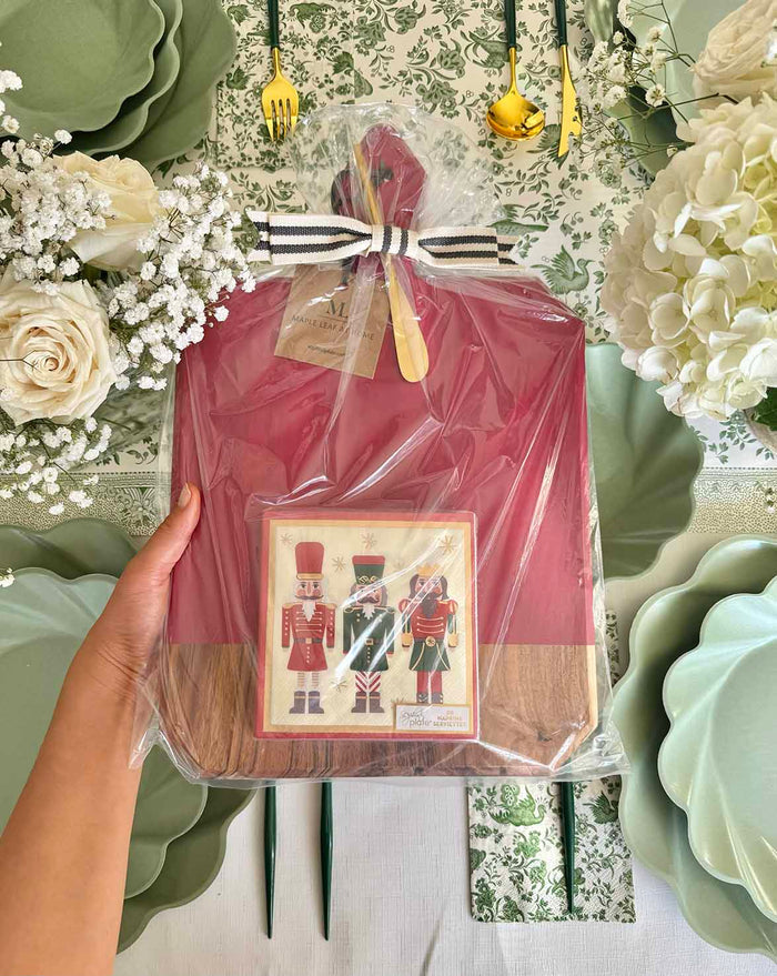 A hand holds the Sophistiplate Christmas Board Gift Kit: Nutcracker, featuring an acacia wood utensil, above a festive table set with green plates, floral decor, and white roses.