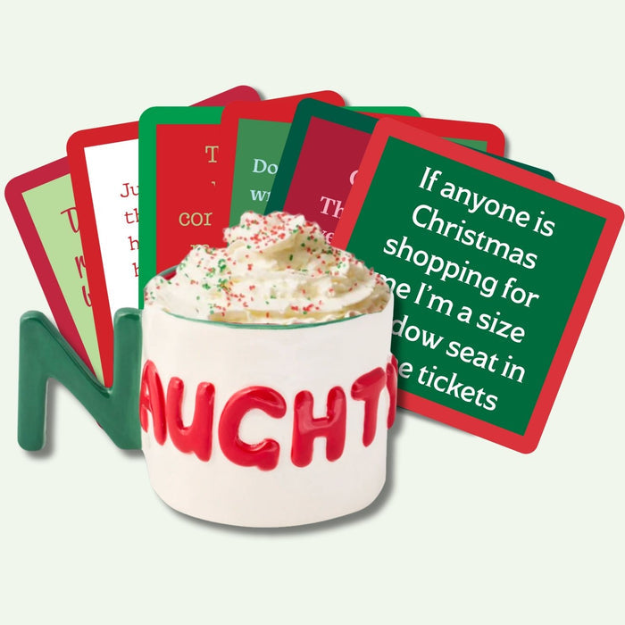 A white mug with "NAUGHTY" in red holds whipped cream with sprinkles. Behind it are red and green coasters from the Drinks On Me Christmas Coaster Bundle: Version 2, including one with a funny holiday travel quote—ideal hostess gifts or stocking stuffers.