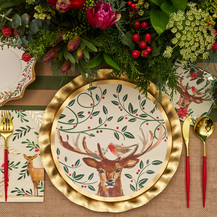 A festive table setting features a Sophistiplate Christmas Forest Dinner Plate from the 8-pack collection, adorned with a charming deer and bird illustration, paired with a gold charger, red-handled cutlery, a floral napkin, and accented by a vibrant flower and foliage centerpiece.