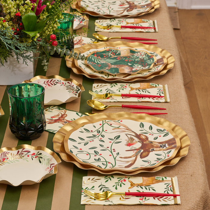 Sophistiplate’s Christmas Forest Dinner Plates (8pk) set a festive table with their gold-edged rims and elegant bird and foliage motifs, complemented by gold cutlery with red handles, a green glass, and a floral centerpiece arranged on a burlap and green table runner.