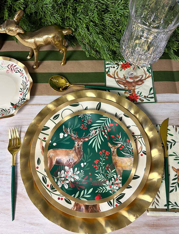 A festive table setting featuring gold utensils, a glass, and Sophistiplate’s Christmas Forest Salad Plates (8pk), displaying deer and foliage on a green background. The décor is completed with evergreen branches and a gold deer figurine.