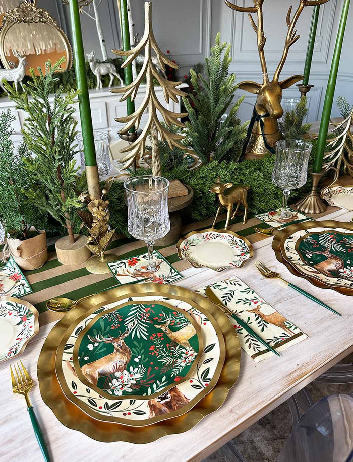 A festive holiday table is set with Sophistiplate’s Christmas Forest Salad Plates (8-pack), gold plates, wavy edge plates, gold utensils, crystal glasses, and green and gold Christmas decorations—including tree and reindeer figurines—arranged on a light wood table.