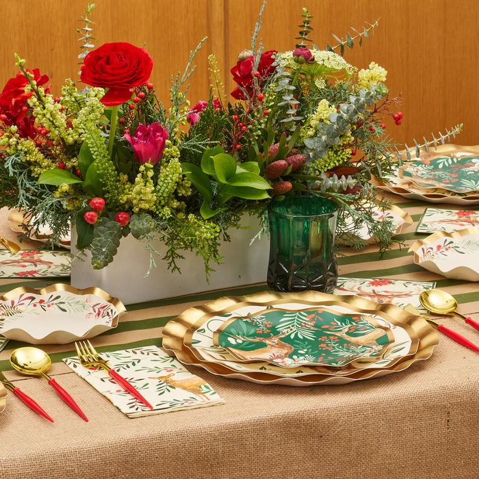 A festive table setting featuring Sophistiplate’s Christmas Forest Salad Plates with elegant gold-rimmed wavy edges, paired with gold cutlery, red-handled forks, floral napkins, and a green glass—all arranged around a vibrant flower centerpiece in a white vase atop a burlap table runner.