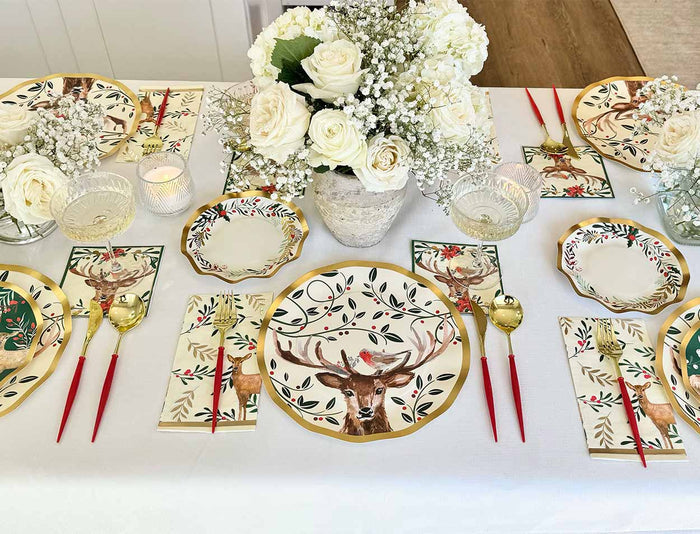 The Sophistiplate Christmas Forest Table Setting features disposable plates, napkins, and cups with deer and holiday foliage designs, red-handled cutlery, gold heart spoons, and a white rose centerpiece in glass vases.