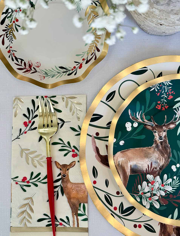 Create a festive table with the Christmas Forest Table Setting by Sophistiplate, featuring disposable plates and napkins with deer and botanical designs, plus a gold fork with red handle. White flowers accent the scene.