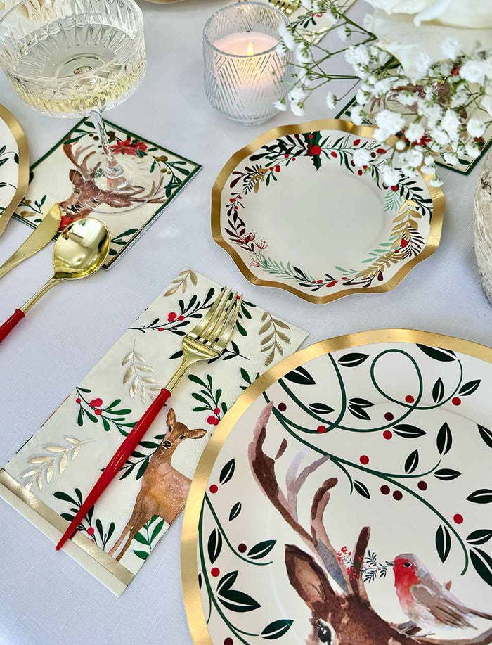 The Christmas Forest Table Setting by Sophistiplate features gold-rimmed disposable plates and utensils with reindeer, robin, holly, and berry motifs—plus a candle, napkin, and flowers for a festive holiday table.