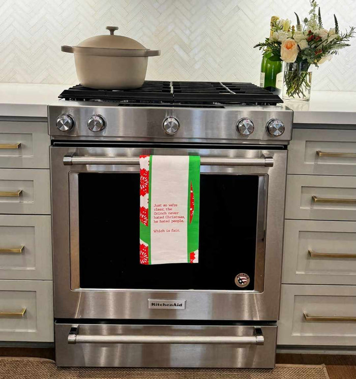 A stainless steel KitchenAid oven with a beige pot sits below the CHRISTMAS Grinch Dish Towel by Drinks On Me, featuring red, green, and white colors and a funny quote—an ideal gift for friends. A vase of flowers is on the right counter.