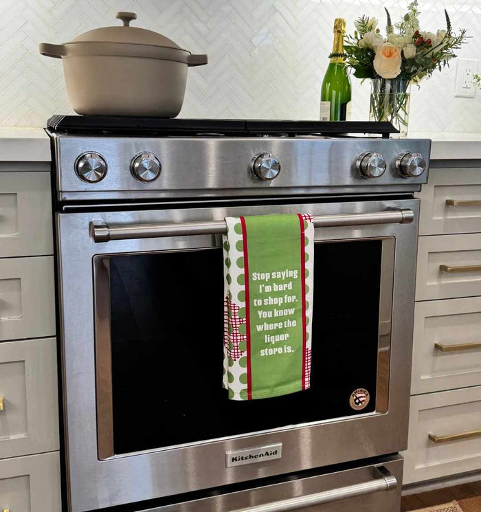 A modern stainless steel oven displays the CHRISTMAS Hard to shop Dish Towel by Drinks On Me, featuring the quote “Stop saying I’m hard to shop for. You know where the liquor store is.” A pot sits on the stove; flowers and champagne are nearby.
