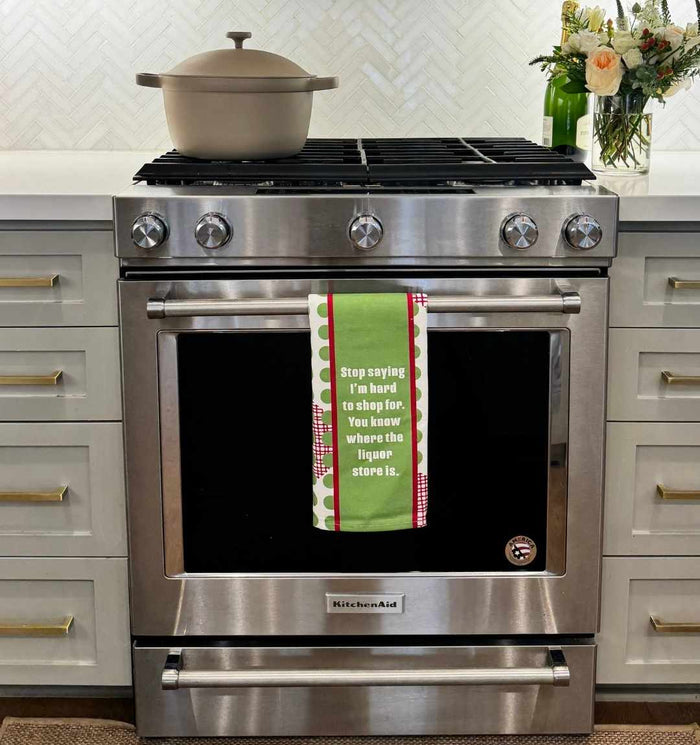 A stainless steel stove features a beige pot and a CHRISTMAS Hard to shop Dish Towel by Drinks On Me, which reads: "Stop saying I'm hard to shop for. You know where the liquor store is." Flowers decorate the counter.
