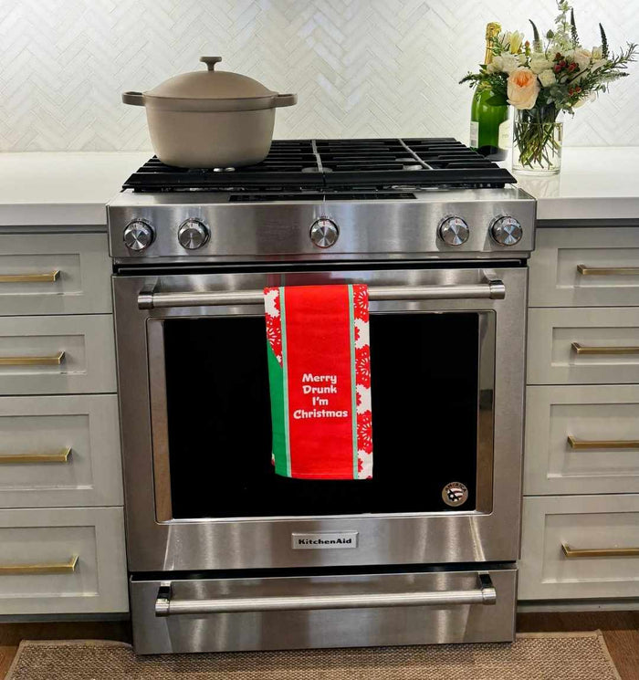 A stainless steel KitchenAid oven displays a beige pot and the Drinks On Me CHRISTMAS Merry Drunk Dish Towel, which reads “Merry Drunk Im Christmas.” A vase of flowers and a bottle sit on the nearby counter.