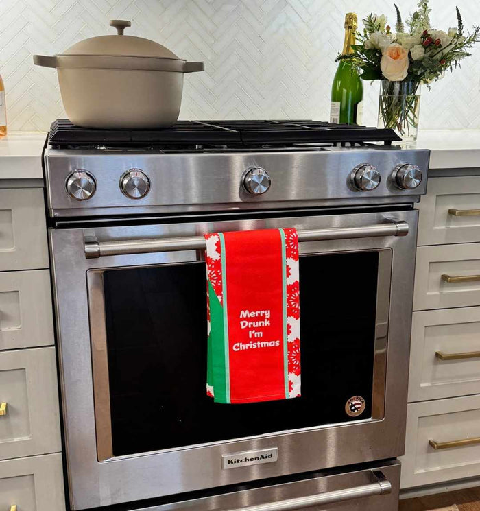 A stainless steel oven displays the CHRISTMAS Merry Drunk Dish Towel by Drinks On Me in red, green, and white, reading "Merry Drunk I’m Christmas." A beige pot sits on the stove with flowers and a gold-topped bottle nearby.